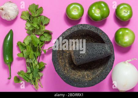 Raw salsa verde ingredients, Mexican food. Tomatillos, onion, garlic, chili peppers and cilantro crushed in a molcajete. Green ingredients on pink Stock Photo
