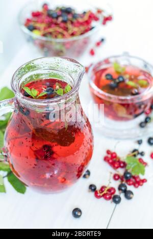 Red and black currant fruit drink in transparent glass cup. Clear glass vase with red and black currant berries on the white wooden background. Stock Photo