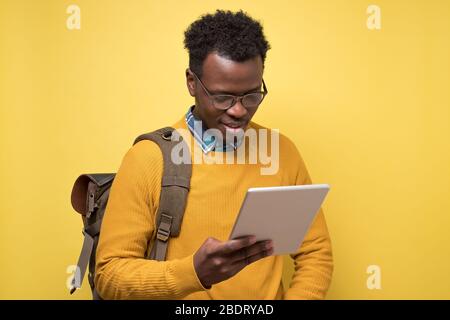 Happy african american college student in glasses holding tablet on yellow wall. Stock Photo
