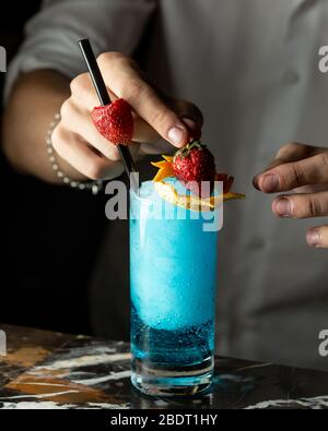 bartender prepares blue cocktail garnished with orange zest and strawberries Stock Photo