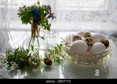 Forest primroses in a retro glass bottle and a crystal vase with eggs on a glass surface . Stock Photo