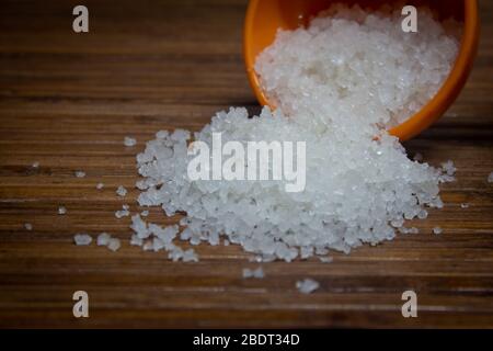 View of the rock salt crystals falling from a measuring cup over a brown wooden background Stock Photo
