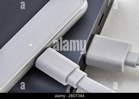 The smartphone is charging from external powerbank with cable. Charge indicator is on. White background, closeup, selective focus Stock Photo