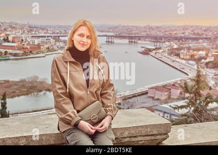 Young smiling beautiful caucasian woman in casual raincoat at the viewpoint of Pierre Loti in Istanbul, Eyup area, she is sitting on the stone with ci Stock Photo