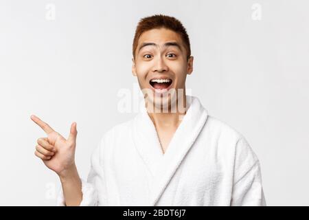 Beauty, spa and leisure concept. Excited, happy asian man in bathrobe laughing and smiling joyfully, pointing finger left, showing way to Stock Photo
