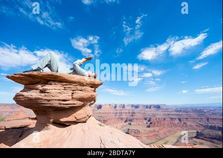 Office worker reclining outdoors with a tablet on mesa rock above a dramatic canyon landscape Stock Photo