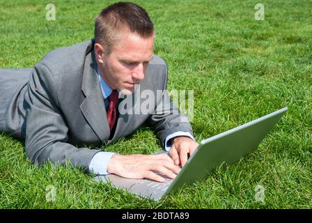 Businessman lying on a sunny patch of green summer grass typing on his laptop computer Stock Photo