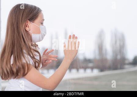 A sad girl in a protective mask from viruses on her face sits by the window.Coronavirus pneumonia COVID-19.The epidemic of coronavirus worldwide. Stock Photo