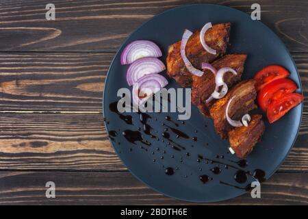 Fried pork belly with sauce and onions in a dark gray plate on a wooden tabletop Stock Photo