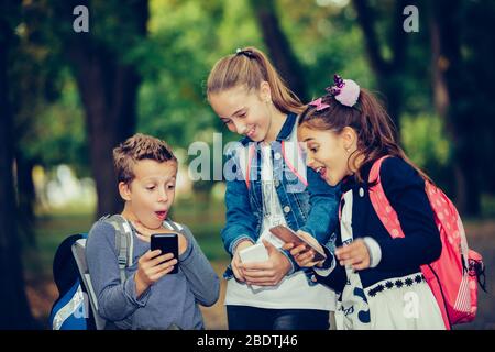 Group of smiling friends hang out after school and using smart phone while walking through the park. Kids laughing and having fun with phone. Selectiv Stock Photo