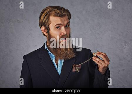 It's time! Yong handsome bearded man looking at his pocket watch while isolated grey wall background. Stock Photo