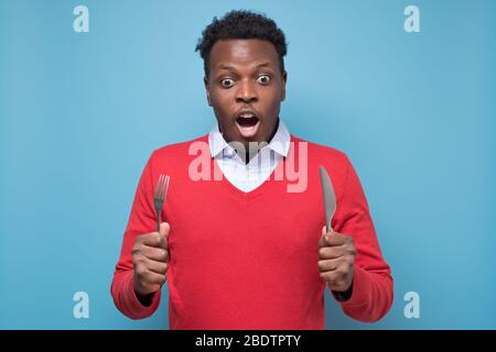 Young african american man with a knife and a fork is shocked with tasty meal. Stock Photo