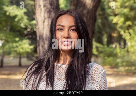 Portrait of happy Indian young woman smiling looking at camera  with dark long hair isolated green trees park background Stock Photo