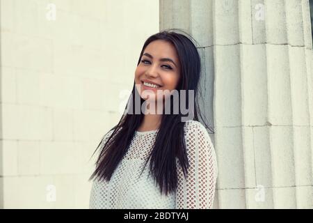 Closeup portrait of confident smiling happy pretty indian young woman in white dress, isolated white building column wall background. Positive human e Stock Photo