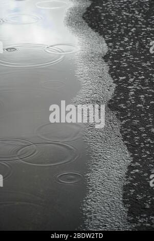 Where an edge of a puddle meets a black asphalt path, raindrops fall into the water and onto the asphalt, creating a surreal abstract of many textures Stock Photo