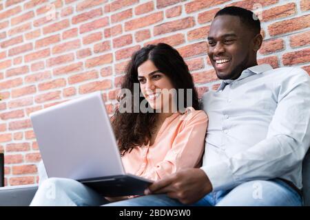 Portrait Of A Happy Young Couple Sitting On Couch Using Laptop Stock Photo