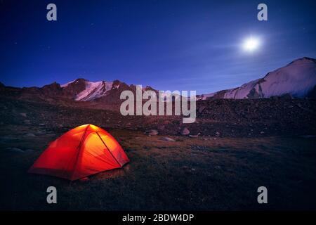 Orange tent glows under night sky full of stars and moon in the mountains in Kazakhstan Stock Photo