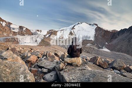 Hiker in brown jacket on stones in front of high snowy mountains at rising moon background Stock Photo