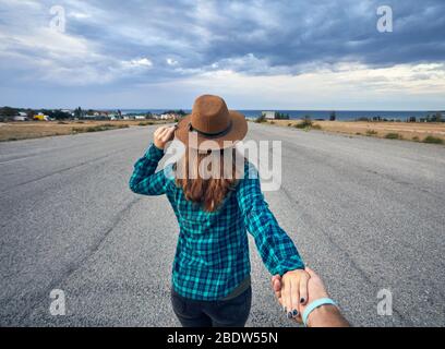 Happy couple in checked shirt holding by hands and walking on the wide asphalt road with lake and cloudy sky background Stock Photo