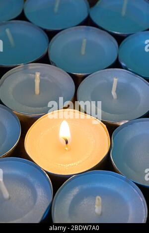 Closeup of tea lights with one being lit. A single warm orange flame between  candles in blue shadow. Christmas, mourning or condolence concept. Stock Photo