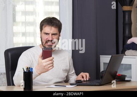 a bearded man is sitting at a table with a laptop in a white sweater looking for a job calling on a mobile phone Stock Photo