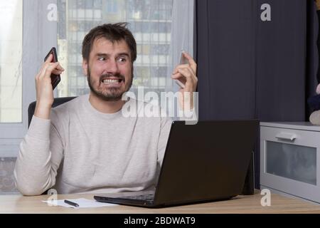 a bearded man is sitting at a table with a laptop in a white sweater looking for a job calling on a mobile phone Stock Photo