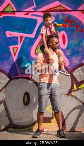 Man with a child on his shoulders standing in front of a graffiti-covered wall at Notting Hill Carnival, London Stock Photo