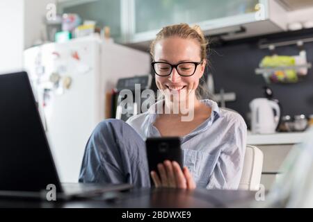 Stay at home and social distancing. Woman in her casual home clothing working remotly from kitchen dining table. Video chatting using social media Stock Photo