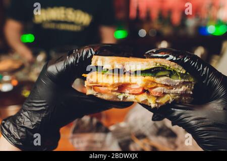 Woman's hands in black rubber gloves are holding juicy black bun burger with meat cutlet, lettuce, tomato, shredded cheese and marinated cucumber. cor Stock Photo