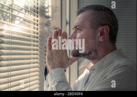 Man in isolation praying near a window blinds. Coronavirus outbreak. Man looks at a sunny street through the window blinds Stock Photo