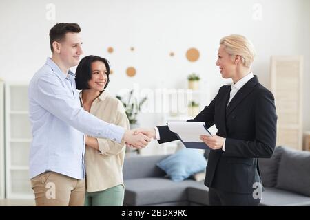 Mature realtor in black suit shaking hands with young couple and congratulating their with purchase of new house Stock Photo