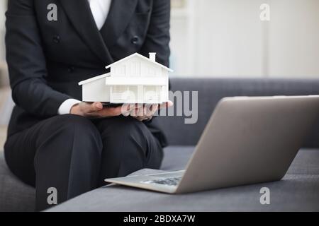 Close-up of real estate agent in black suit sitting on sofa and holding the model of the house in her hands while working online Stock Photo