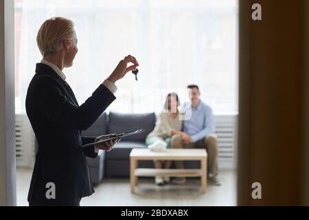 Rear view of realtor in black suit holding keys from the home and showing them to young couple who sitting on sofa in the room Stock Photo