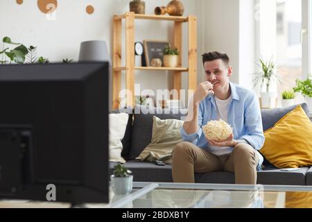 Young man sitting on sofa eating popcorn and watching his favourite film on TV at home Stock Photo