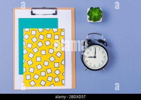 Pile of bright notebooks, clipboard, alarm- clock, table plant and stationary on blue desk top. Copy space. Free space. Start of working day.  Stock Photo