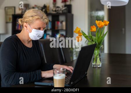 Middle aged Caucasian woman working from home on laptop in her living room, wearing face protective mask whilst being in quarantine and isolation Stock Photo