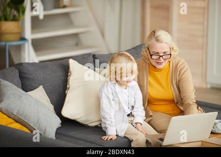 Mature woman in eyeglasses talking to her grandson and using laptop computer while they sitting on sofa in the living room Stock Photo