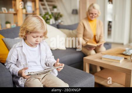 Little boy with blond hair playing computer game on tablet pc with his grandmother sitting on sofa in the background at home Stock Photo
