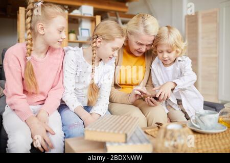 Grandmother sitting on sofa and using her mobile phone with her grandchildren who helping her and teaching her in the room at home Stock Photo