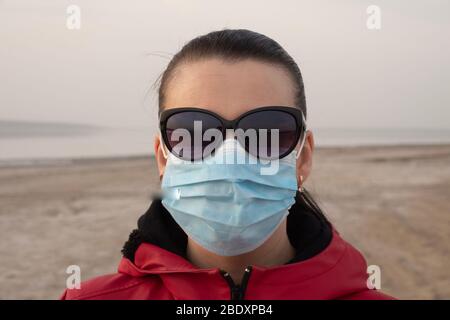 A brunette woman in sunglasses and a red jacket and a protective surgical mask on an empty beach during quarantine. Stock Photo