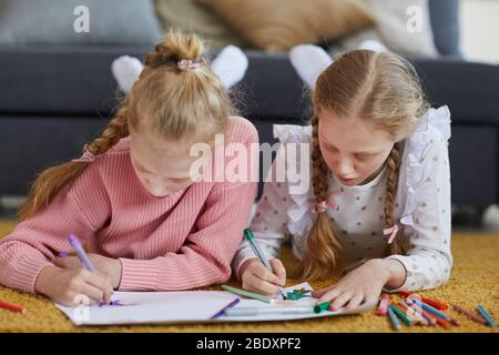 Two little girls lying on the floor and drawing pictures with pencils in the room at home Stock Photo