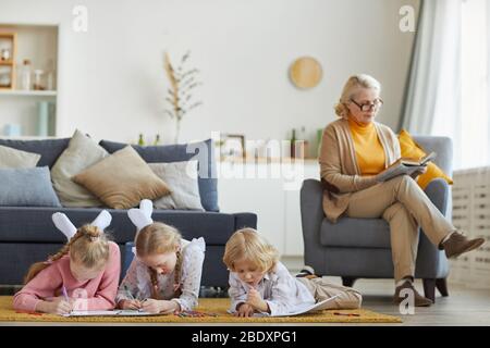 Children lying on the floor and drawing with pencils with their grandmother sitting on armchair and reading a book Stock Photo