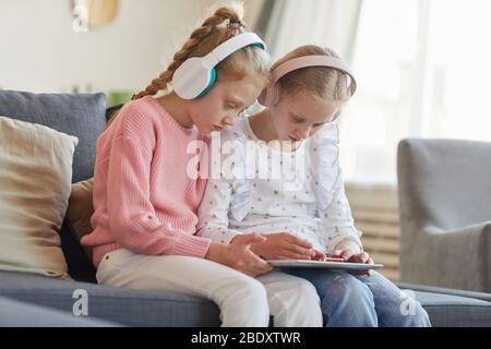 Two girls in headphones sitting on sofa and using digital tablet in their room at home Stock Photo