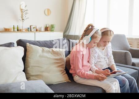 Two sisters in headphones sitting on sofa with digital tablet and playing together in online games at home Stock Photo