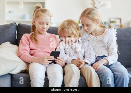 Two sisters sitting on sofa together with their younger brother and using mobile phone to play in games at home Stock Photo