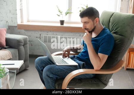 Young focused man studying at home during online courses or free information by hisself. Becomes programmer, designer, marketer while isolated, quarantined. Using laptop, smartphone, headphones. Stock Photo