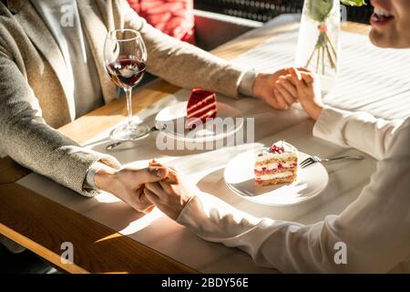Two young romantic dates holding by hands over served table while having tasty cakes and red wine after lunch in classy restaurant Stock Photo