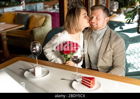 Young happy woman with luxurious bouquet of red roses kissing her boyfriend on cheek while both sitting by table in restaurant Stock Photo