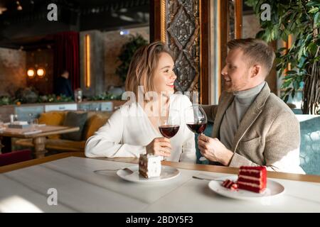 Young romantic couple with glasses of red wine talking and making toast by served table after lunch in luxurious restaurant Stock Photo