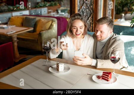Happy young woman pointing at smartphone screen while discussing something with her boyfriend by served table in restaurant Stock Photo